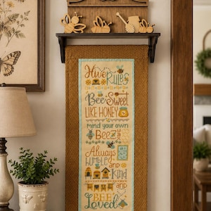 May include: A decorative wall hanging with a bee-themed design. The piece features a framed embroidered panel with the text "Hive Rules, Bee Sweet Like Honey, Mind Your Own Beez, Always Humble and Kind, Bee Loved." Wooden bee cutouts adorn the top.