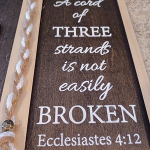 May include: Wooden framed sign with the text "A cord of three strands is not easily broken" from Ecclesiastes 4:12. The sign has a dark brown background with white lettering and a braided rope detail.