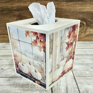 May include: A square tissue box with a fall-themed design. The box features a white exterior with a printed image of pumpkins, autumn leaves, and a window scene. White tissues are visible at the top.