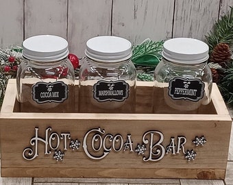 May include: A wooden box with the words "Hot Cocoa Bar" on the front. The box contains three glass jars with white lids. The jars are labeled "Cocoa Mix", "Marshmallows", and "Peppermint".