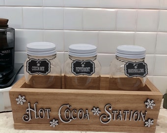 May include: Wooden box with the words "Hot Cocoa Station" on the front. The box contains three glass jars with white lids. The jars are labeled "Cocoa Mix", "Peppermint", and "Marshmallows".