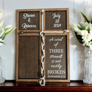 May include: Wooden cross-shaped sign with the names "Steven & Rebecca" and the date "July 28 2024". The sign includes the quote "A cord of THREE strands is not easily BROKEN" from Ecclesiastes 4:12. The sign is decorated with rope.