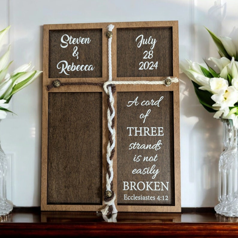 May include: Wooden cross-shaped sign with the names "Steven & Rebecca" and the date "July 28 2024". The sign includes the quote "A cord of THREE strands is not easily BROKEN" from Ecclesiastes 4:12. The sign is decorated with rope.