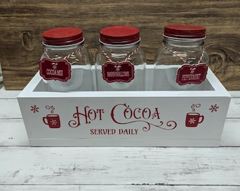 May include: White wooden box with three glass jars with red lids. The jars are labeled "Cocoa Mix", "Marshmallows", and "Peppermint". The box has a sign that says "Hot Cocoa Served Daily" with a red snowflake and a red mug with steam coming out of it.