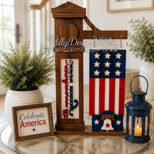 May include: A wooden patriotic decoration featuring a sign that says "America" in red, white, and blue. A framed sign reads "Celebrate America". A blue lantern with a lit candle sits nearby. The decoration has a flag design.