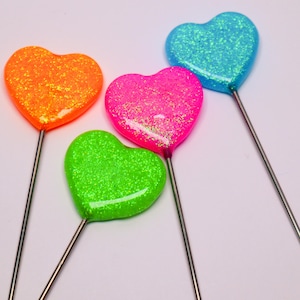 May include: Four heart-shaped decorations on thin metal sticks. Each heart features a vibrant, glittery finish in a distinct neon color: orange, bright pink, lime green, and cyan blue. The hearts have a smooth, glossy, and reflective surface.