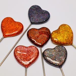 May include: A collection of six heart-shaped decorative picks, each featuring a smooth, glossy finish embedded with vibrant glitter. The hearts come in various colours including orange, dark blue, deep red, gold, and dark grey, attached to thin metal stems.