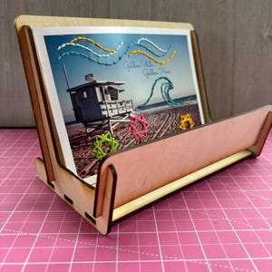 Puede incluir: Un soporte de madera para tarjetas con una postal que muestra una caseta de salvavidas en una playa. La postal tiene detalles bordados de olas y el texto "Golden State Golden Hour".