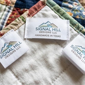 May include: A patchwork quilt featuring a variety of floral and striped fabric squares in shades of blue, red, and tan. Three white rectangular fabric labels are attached, each printed with the Signal Hill Designs LLC logo and text.