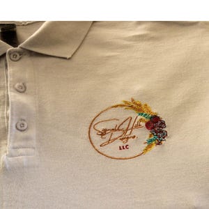 May include: A beige polo shirt with an embroidered logo. The logo is a circle with a wheat stalk and flowers. The text "Signature Hill Designs LLC" is inside the circle.