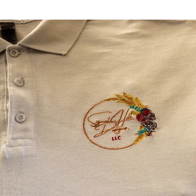 May include: A beige polo shirt with an embroidered logo. The logo is a circle with a wheat stalk and flowers. The text "Signature Hill Designs LLC" is inside the circle.