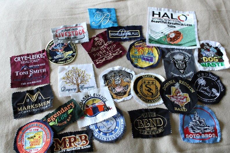 May include: A collection of embroidered patches with various designs and text. Patches include company logos, text like "HALO," "Legacy Hospice," and "Mighty Redskin Band." Colors include blue, red, gold, and black. The patches are of various shapes and sizes.