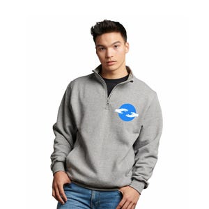 May include: A gray quarter-zip sweatshirt with a blue circle logo featuring two hands reaching towards each other. The sweatshirt has a black undershirt and is worn with blue jeans. The zipper is silver.
