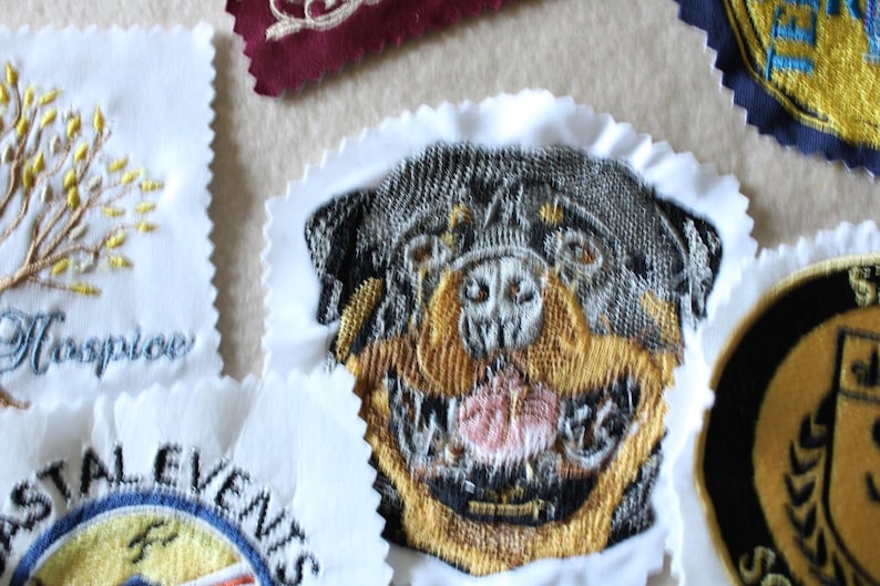 May include: Embroidered patches with various designs, including a Rottweiler dog, a tree, and text. The patches feature detailed stitching in multiple colors, with a white scalloped edge. The patches are likely for clothing or accessories.