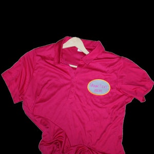 May include: A magenta polo shirt with a three-button placket and short sleeves. The shirt has a light blue oval patch with the text "YOUR TEXT HERE" embroidered in a cursive font. The shirt is hanging on a wooden hanger.