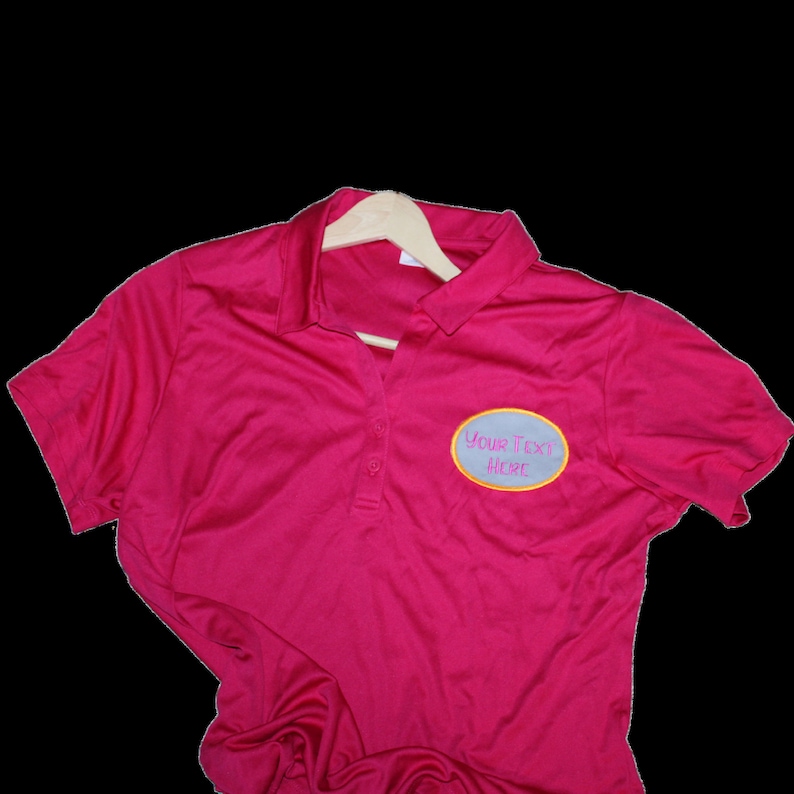 May include: A magenta polo shirt with a three-button placket and short sleeves. The shirt has a light blue oval patch with the text "YOUR TEXT HERE" embroidered in a cursive font. The shirt is hanging on a wooden hanger.