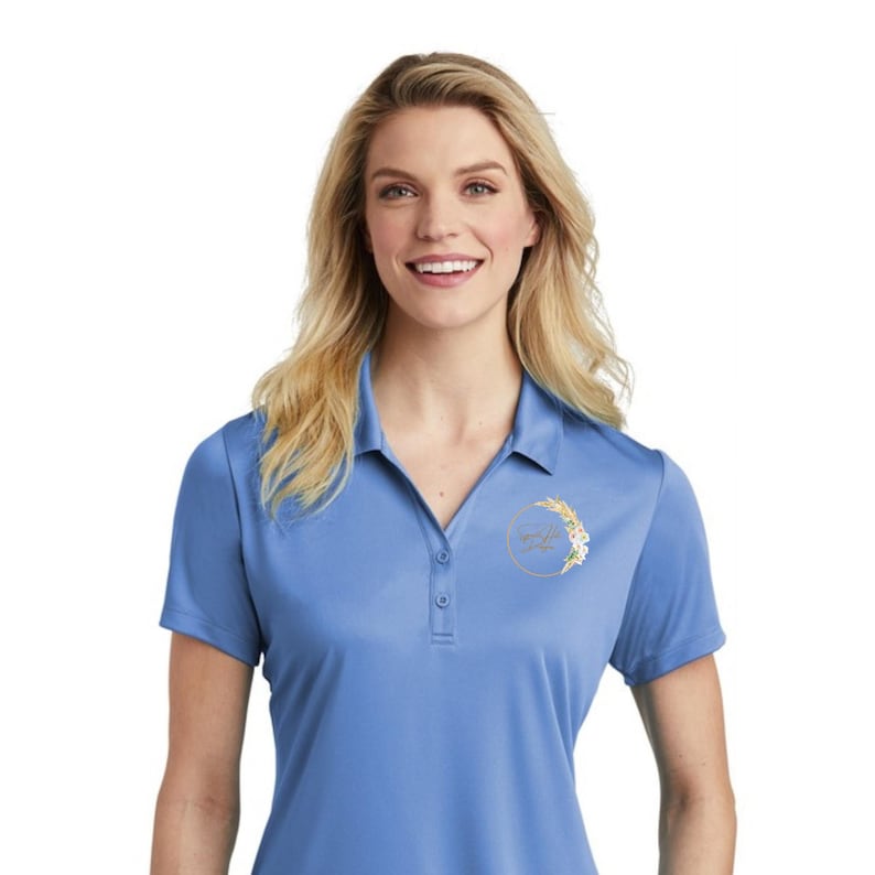 May include: A light blue polo shirt with short sleeves and a collared neckline. The shirt features a three-button placket and a floral design inside a gold circle on the chest. The model is smiling.