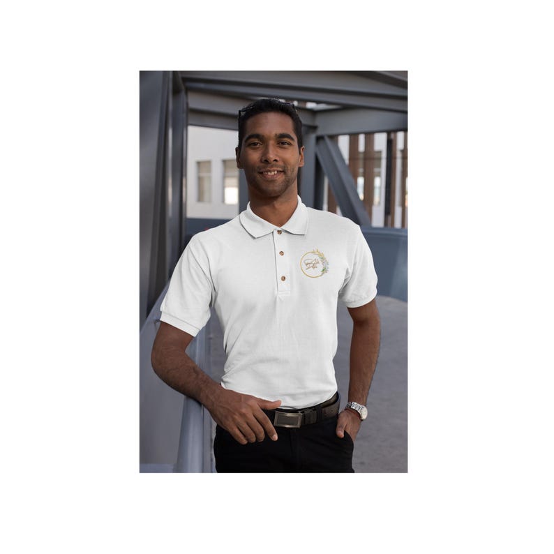 May include: A white polo shirt with a gold embroidered logo on the left chest. The logo is a circle with a floral design and the text "Simply Inspired" inside.