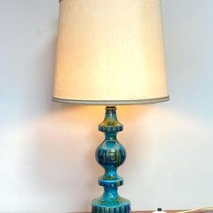 May include: A vintage table lamp with a cream-colored lampshade and a turquoise ceramic base. The base has a bulbous shape with a glossy finish and green accents. The lamp is plugged in and turned on.