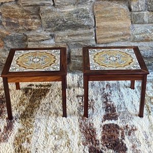 May include: Two matching square side tables with dark wood frames and decorative tile tops. The tiles feature a gold, brown, and white geometric design. The tables are set on a shaggy, multi-colored rug.
