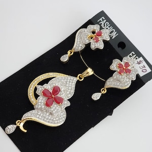 Könnte beinhalten: Ein goldener Anhänger-Halsketten- und Ohrring-Set mit einem roten Blumendesign und klaren Steinen. Der Anhänger ist in Form eines gebogenen Blattes mit einer roten Blume in der Mitte. Die Ohrringe sind klein und haben ein rotes Blumendesign mit klaren Steinen. Das Set ist mit "Fashion Ear 30 €" gekennzeichnet.