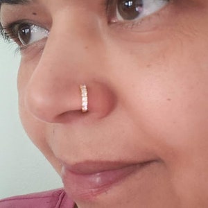 Puede incluir: Primer plano de la cara de una persona con un peque&ntilde;o aro de nariz dorado con brillantes transparentes. El aro de nariz es un dise&ntilde;o simple y elegante.
