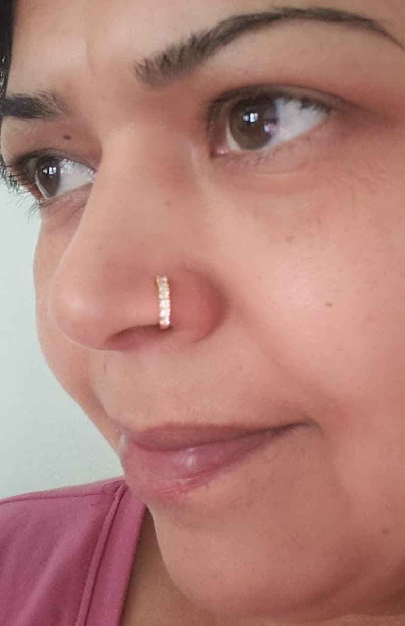 Puede incluir: Primer plano de la cara de una persona con un peque&ntilde;o aro de nariz dorado con brillantes transparentes. El aro de nariz es un dise&ntilde;o simple y elegante.