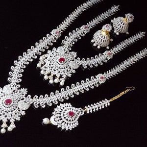Pode incluir: Um conjunto de joias de noiva indianas tradicionais que inclui um colar longo de várias voltas com um grande pingente adornado, brincos combinando e um maang tikka. As joias são feitas de prata e são adornadas com pedras brancas brilhantes e detalhes vermelhos.