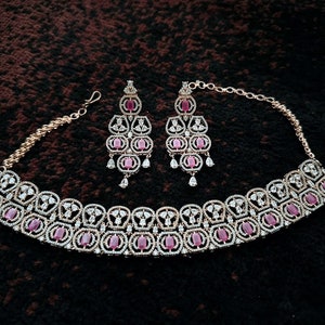 Pode incluir: Um colar choker de ouro com uma fileira de pedras cor-de-rosa dispostas em um padrão geométrico. Brincos combinando com pedras cor-de-rosa pendurados em ganchos de ouro.