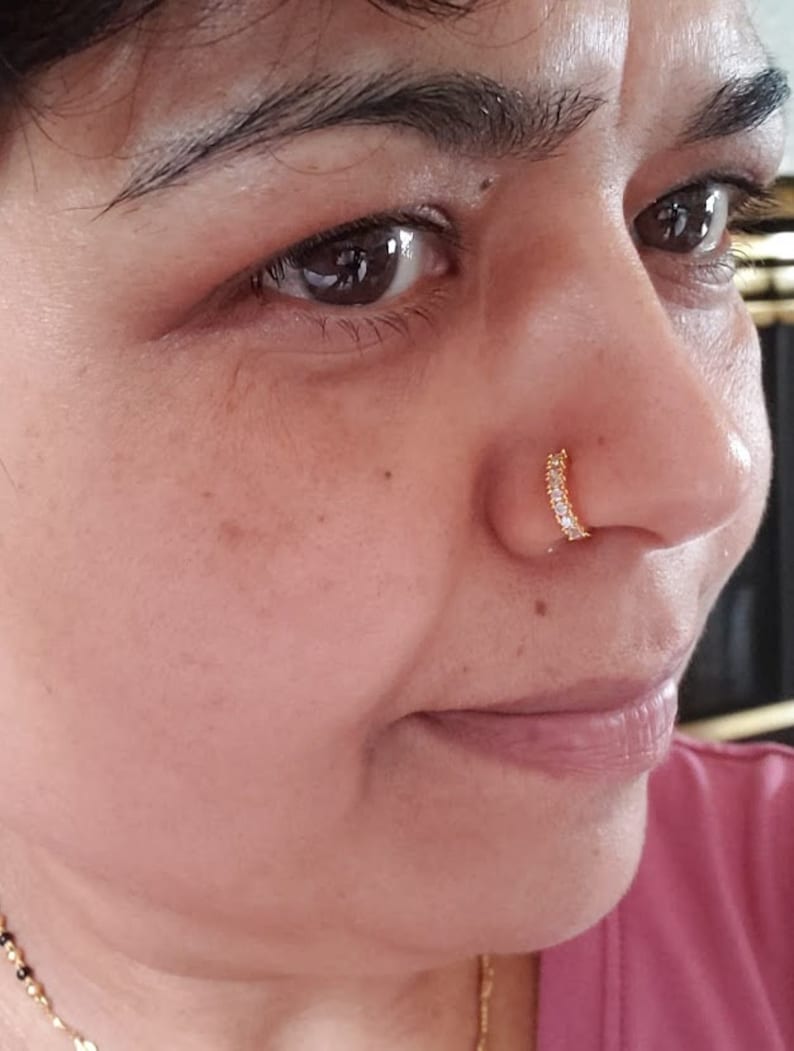 Puede incluir: Primer plano de la cara de una persona con un peque&ntilde;o aro de nariz de oro con piedras transparentes. El aro de nariz es de estilo aro.