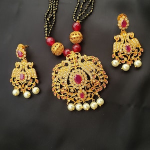 May include: A gold necklace with a peacock design pendant and red and white stones. The necklace is on a black string and has a pearl drop. The set also includes matching earrings with a peacock design and red and white stones.