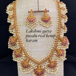 Puede incluir: Un collar de oro con un colgante de Lakshmi y detalles de perlas. El collar está formado por varias hileras de perlas y cuentas doradas. El colgante presenta un diseño detallado de Lakshmi, la diosa hindú de la riqueza y la prosperidad. El collar viene acompañado de un par de pendientes a juego con un diseño similar.