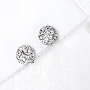 May include: A pair of silver cufflinks with a circular design featuring a swirl pattern and clear rhinestones.