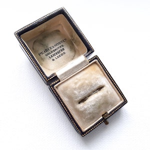 May include: An open black ring box with gold trim and a white velvet interior. The box is labelled "Pearce & Sons Ltd. Goldsmiths, Leicester & Leeds."
