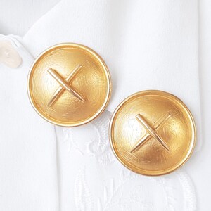 May include: A pair of gold-tone earrings with a textured, circular design. Each earring features a raised, crossed-line design in the centre.