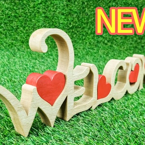 May include: Wooden "WELCOME" sign with red heart accents. The letters are light brown, with red hearts integrated into the design. The word "NEW" is displayed in a red and yellow box. The sign is placed on a green, grassy surface.