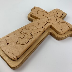 May include: A wooden puzzle in the shape of a cross. The puzzle is made of light-colored wood and features animal shapes within the cross design. The pieces are layered, creating a three-dimensional effect. The cross is set against a white background.
