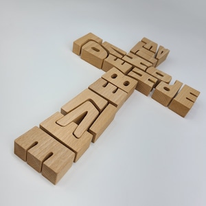 Puede incluir: Rompecabezas de madera en forma de cruz, con la palabra "ESPERANZA" en la viga vertical y "FE" en la viga horizontal. Las piezas del rompecabezas son de color marrón claro y están sobre una superficie blanca. Las letras están talladas en los bloques de madera.