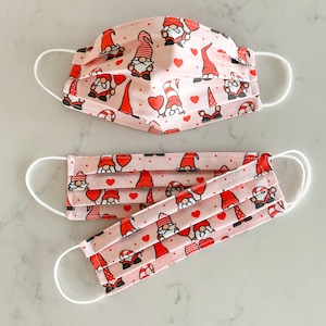 May include: Two pink face masks with white elastic ear loops. Both masks feature a repeating pattern of red hearts and cartoon gnomes.