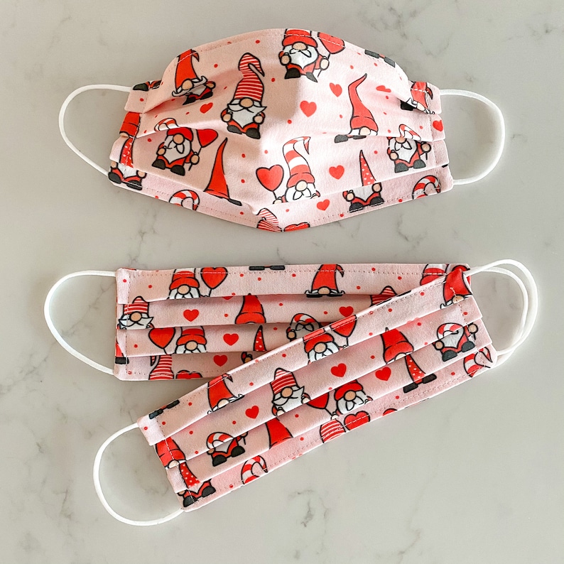 May include: Two pink face masks with white elastic ear loops. Both masks feature a repeating pattern of red hearts and cartoon gnomes.