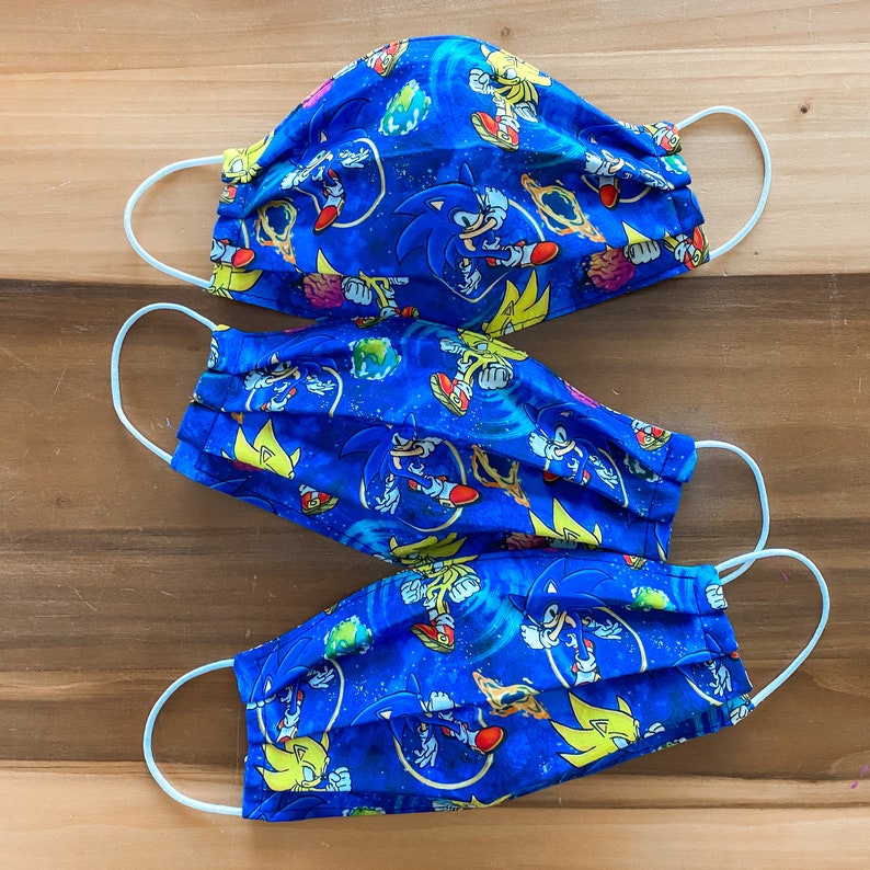 Op de afbeelding: Drie blauwe stoffen mondmaskers met een witte achtergrond en een herhalend patroon van Sonic the Hedgehog-personages. De maskers zijn gemaakt van katoen en hebben elastische oorlussen.