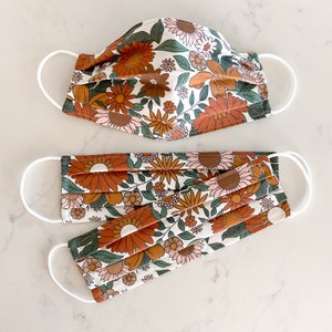 Peut inclure: Deux masques faciaux avec un motif floral dans des tons d'orange, de brun et de blanc. Les masques sont en tissu et ont des boucles d'oreille &eacute;lastiques blanches.