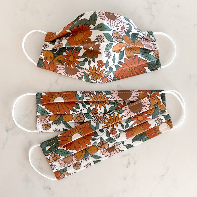 Peut inclure: Deux masques faciaux avec un motif floral dans des tons d'orange, de brun et de blanc. Les masques sont en tissu et ont des boucles d'oreille &eacute;lastiques blanches.
