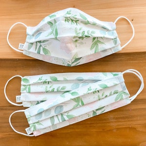 May include: Two white face masks with a floral pattern featuring green leaves and pink flowers. The masks are pleated and have elastic ear loops.