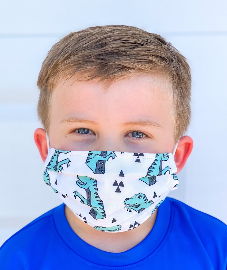 May include: A white face mask with a repeating pattern of green dinosaurs and black triangles. The mask is pleated and has elastic ear loops.