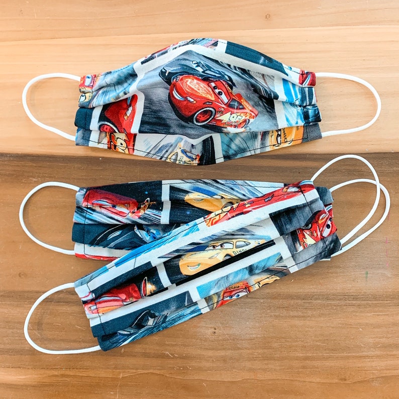 Puede incluir: Dos mascarillas de tela blanca con tem&aacute;tica de Cars. Las mascarillas presentan un fondo azul, negro y blanco con coches de carreras rojos. Las mascarillas son plisadas y tienen lazos el&aacute;sticos blancos para las orejas.