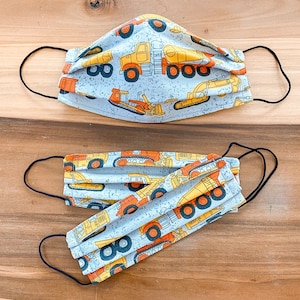 May include: Two face masks with a blue background and a white pattern of construction vehicles. The masks are made of fabric and have elastic ear loops.