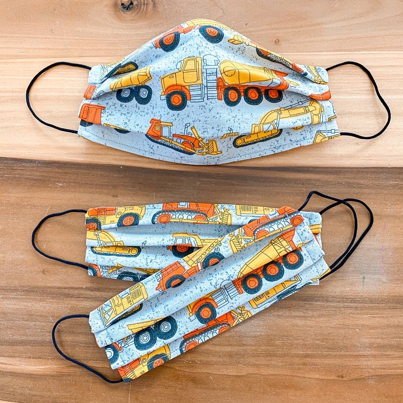 May include: Two face masks with a blue background and a white pattern of construction vehicles. The masks are made of fabric and have elastic ear loops.