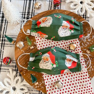 May include: Two green fabric face masks with a white polka dot background. The masks feature a repeating pattern of Santa Claus with a red hat and white beard. The masks are on a wooden surface with white and black checkered fabric and a white polka dot fabric.