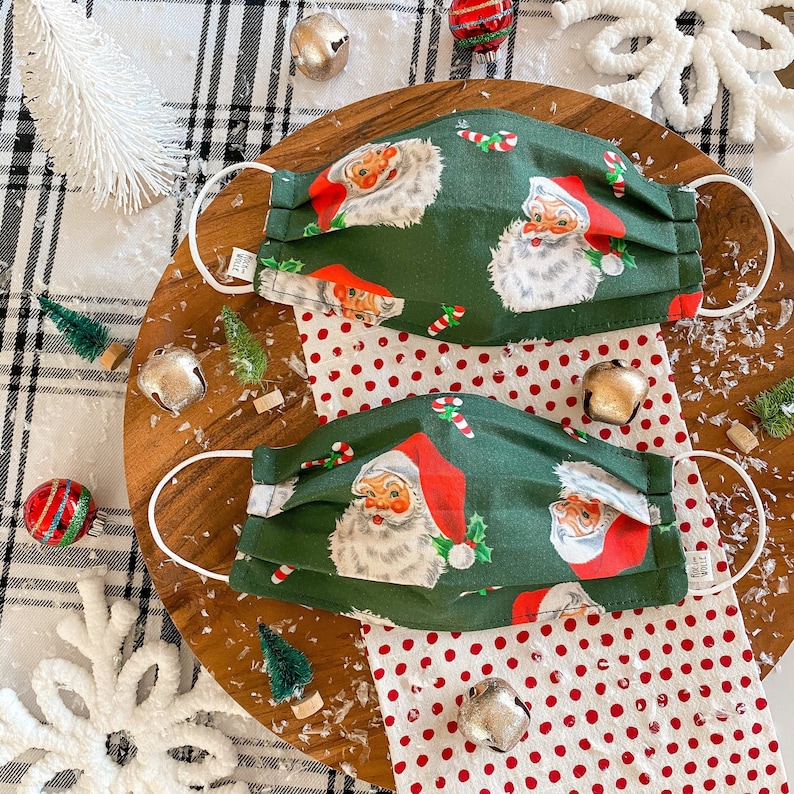 May include: Two green fabric face masks with a white polka dot background. The masks feature a repeating pattern of Santa Claus with a red hat and white beard. The masks are on a wooden surface with white and black checkered fabric and a white polka dot fabric.